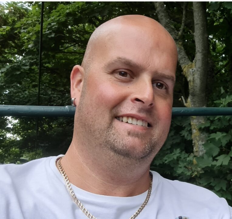 A photo of Steven wearing a white T-shirt and smiling at the camera, with trees in the background.