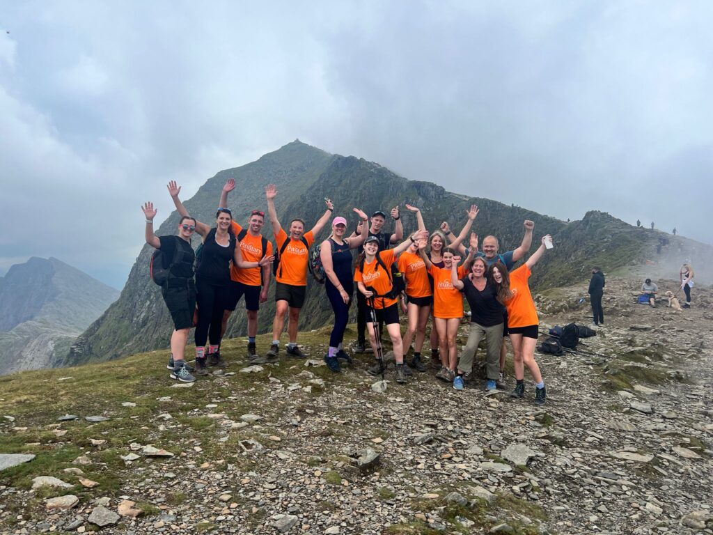 A group of people wearing orange t-shirts and their arms in the air, stood in front of Snowdon.
