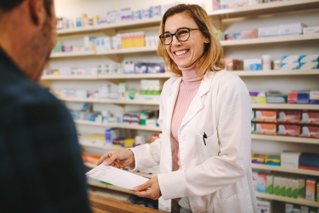 A female pharmacist, wearing a white lab coat, is stood at a medicine counter smiling at a customer
