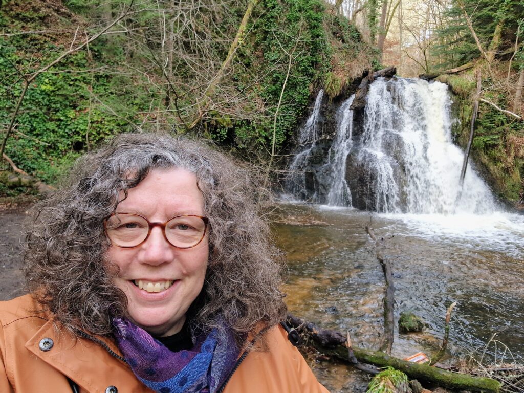 A photo of Ann stood in front of a waterfall. She is wearing an orange raincoat and a colourful scarf