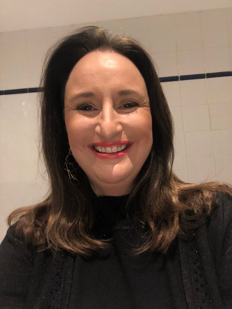 Debbie is wearing a black jumper and has dark hair. She is stood in front of a white wall and is smiling at the camera