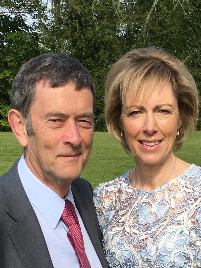 John is stood next to his wife, Cathy. He is wearing a grey suit and she is wearing a pale blue dress