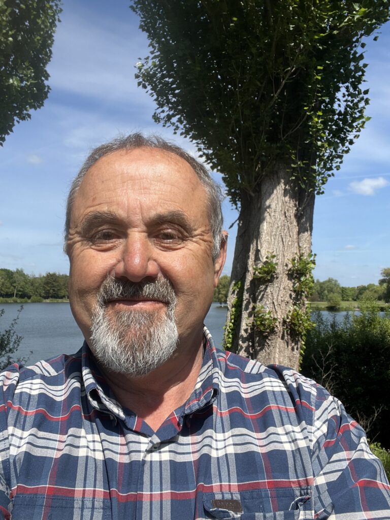 A photo of Phil in front of a lake on a sunny day. He is wearing a blue chequered shirt.