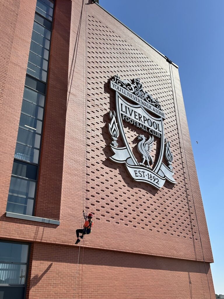 Roz is pictured abseiling down the side of Anfield football stadium. The Liverpool logo is seen on the wall and Roz is hanging from a rope next to it.