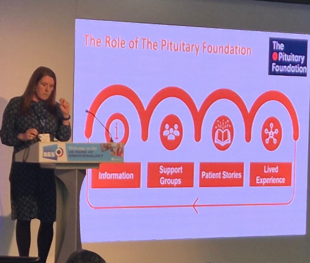 Roz is pictured stood behind a podium, delivering a presentation about her experience as a patient. There is a slide shown behind her that explains the role of The Pituitary Foundation in supporting patients