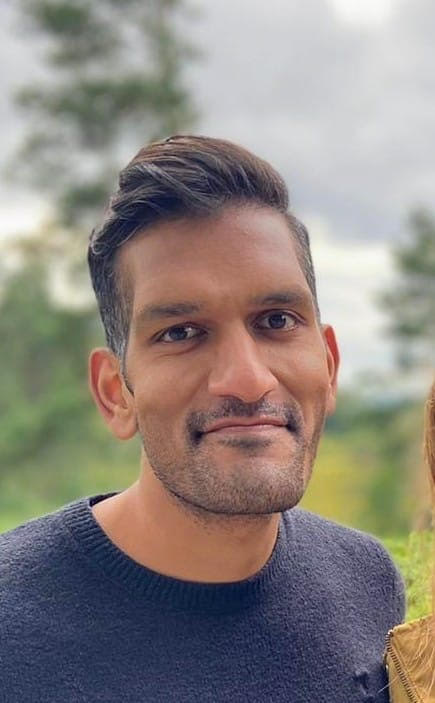Ketan is smiling for a photo in front of some trees. He is wearing a navy blue jumper
