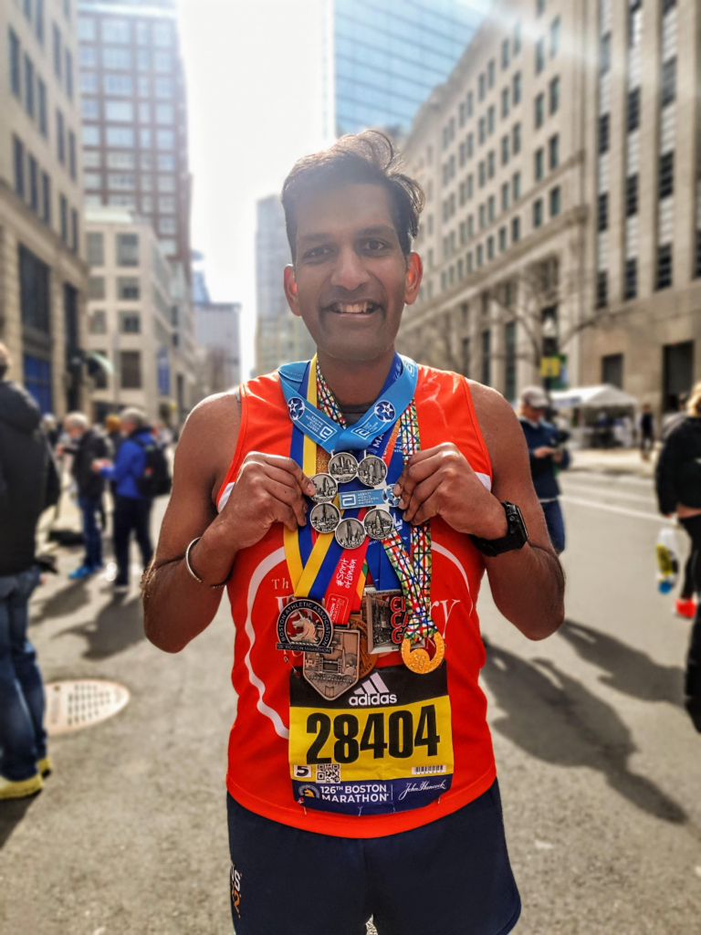 Ketan wearing a PF running top and wearing lots of medals. He has completed a race and is smiling at the camera.