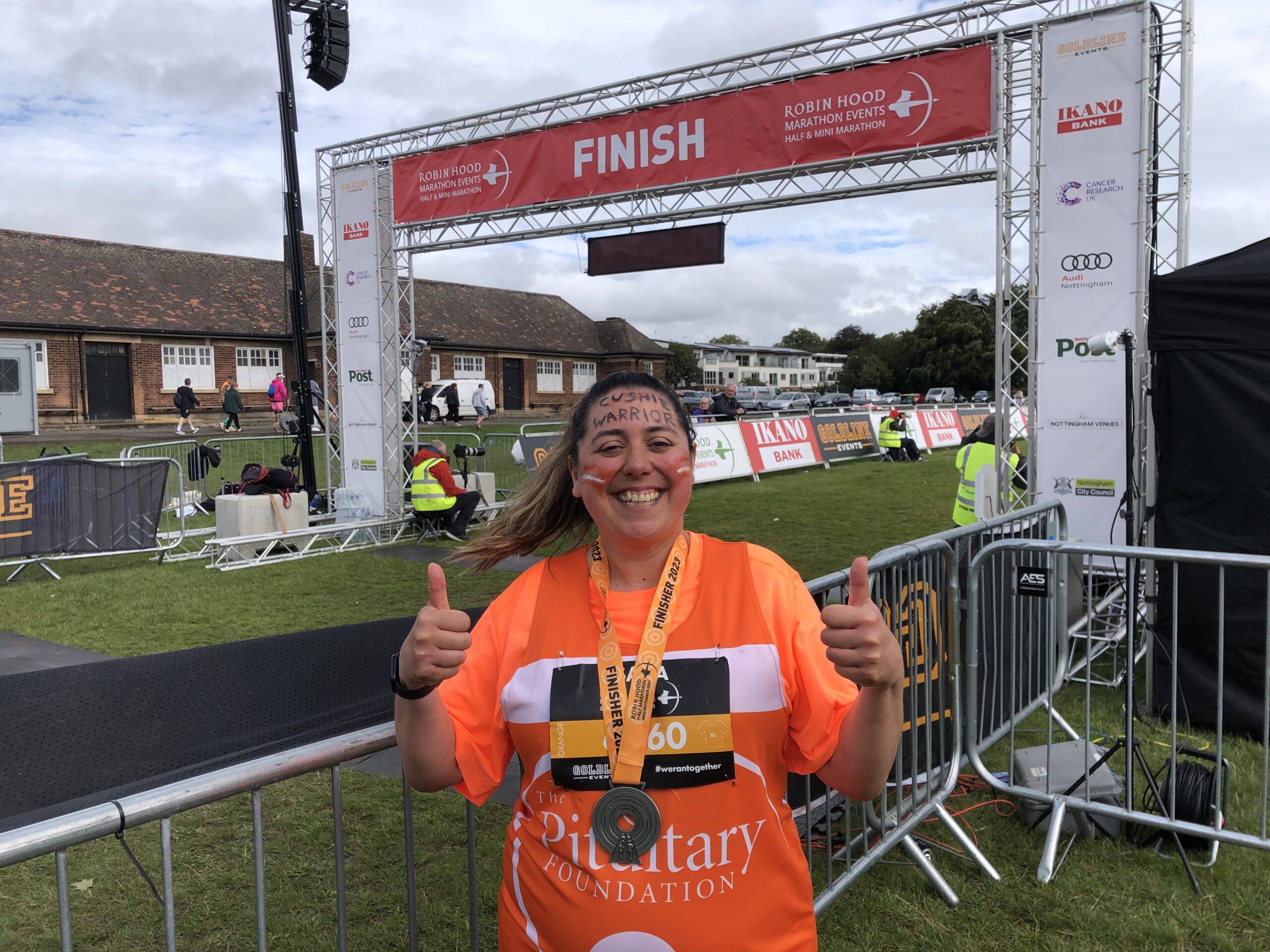 A smiling woman is stood in front of a finish line for a race. She is wearing a bright orange Pituitary Foundation top and is holding her thumbs up