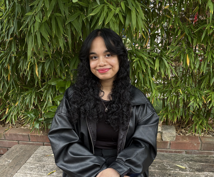 Amber, a young woman with dark hair, is sat outside in front of greenery. She is wearing a black top and black jacket.