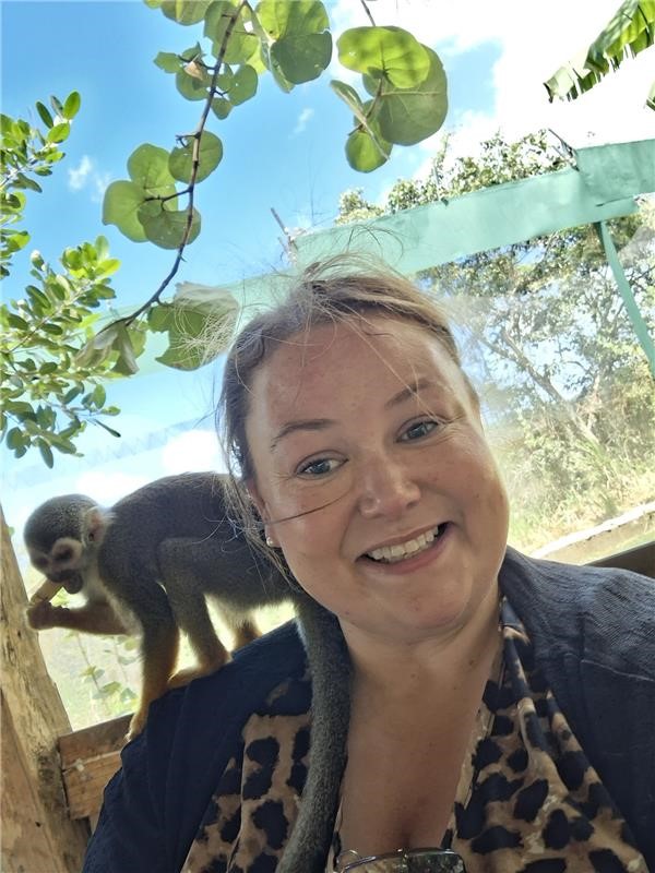 A woman is smiling for a photo. There is a small capuchin monkey on her shoulder. She is outdoors, surrounded by trees and the sun is shining