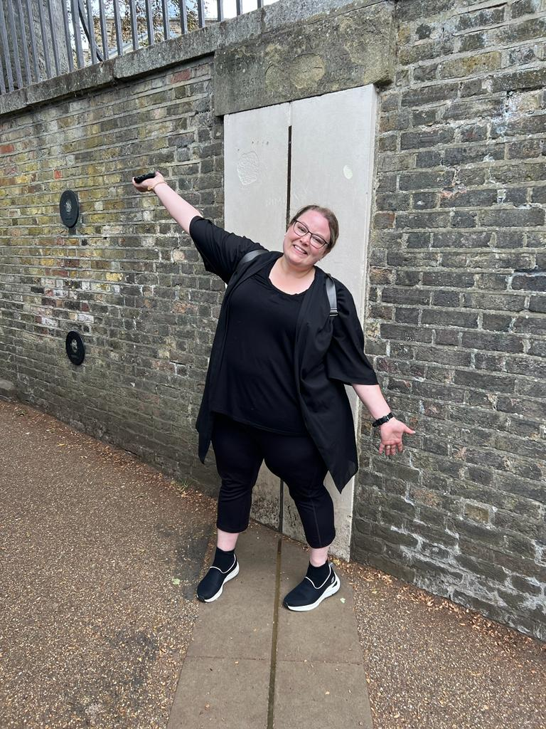 A woman wearing all black is stood in front of a stone wall, she is smiling and holding her arms out