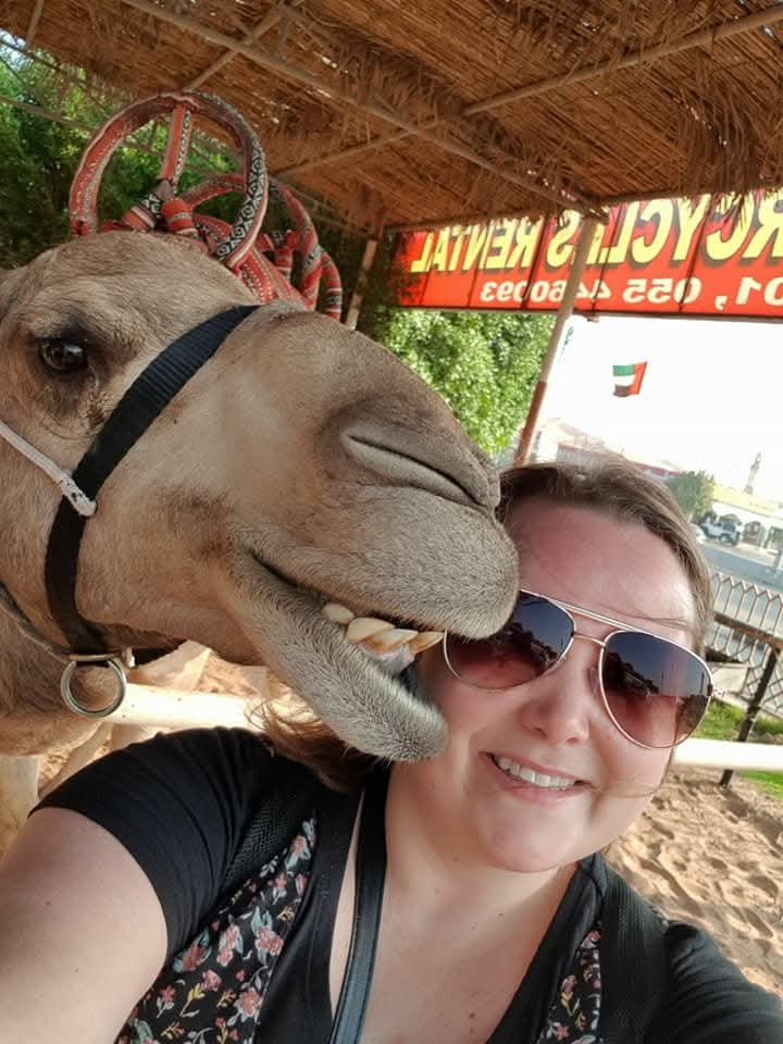 Heather is wearing sunglasses and posing for a photograph stood alongside a camel. The camel looks like it's trying to eat her sunglasses!