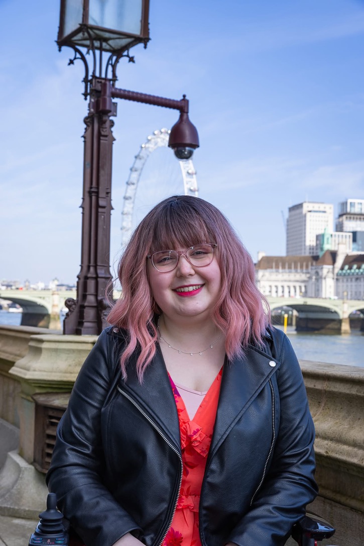 A young woman in London, stood on the bank of the Thames in front of the London Eye on a sunny day. She has pink hair and is wearing red lipstick and a black jacket.