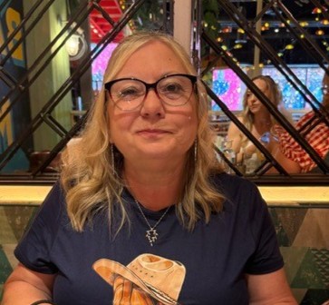 A woman with blond hair and glasses is sat in a restaurant, smiling at the camera