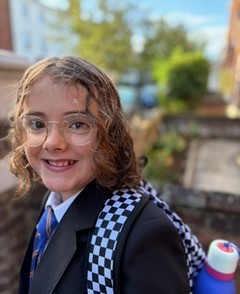 A young girl wearing a school blazer and backpack is smiling at the camera
