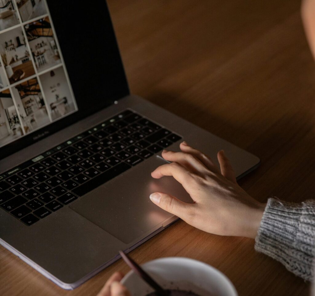 A laptop placed on a table, with a person's hand on the mouse pad. The person is also holding a cup of tea in their other hand