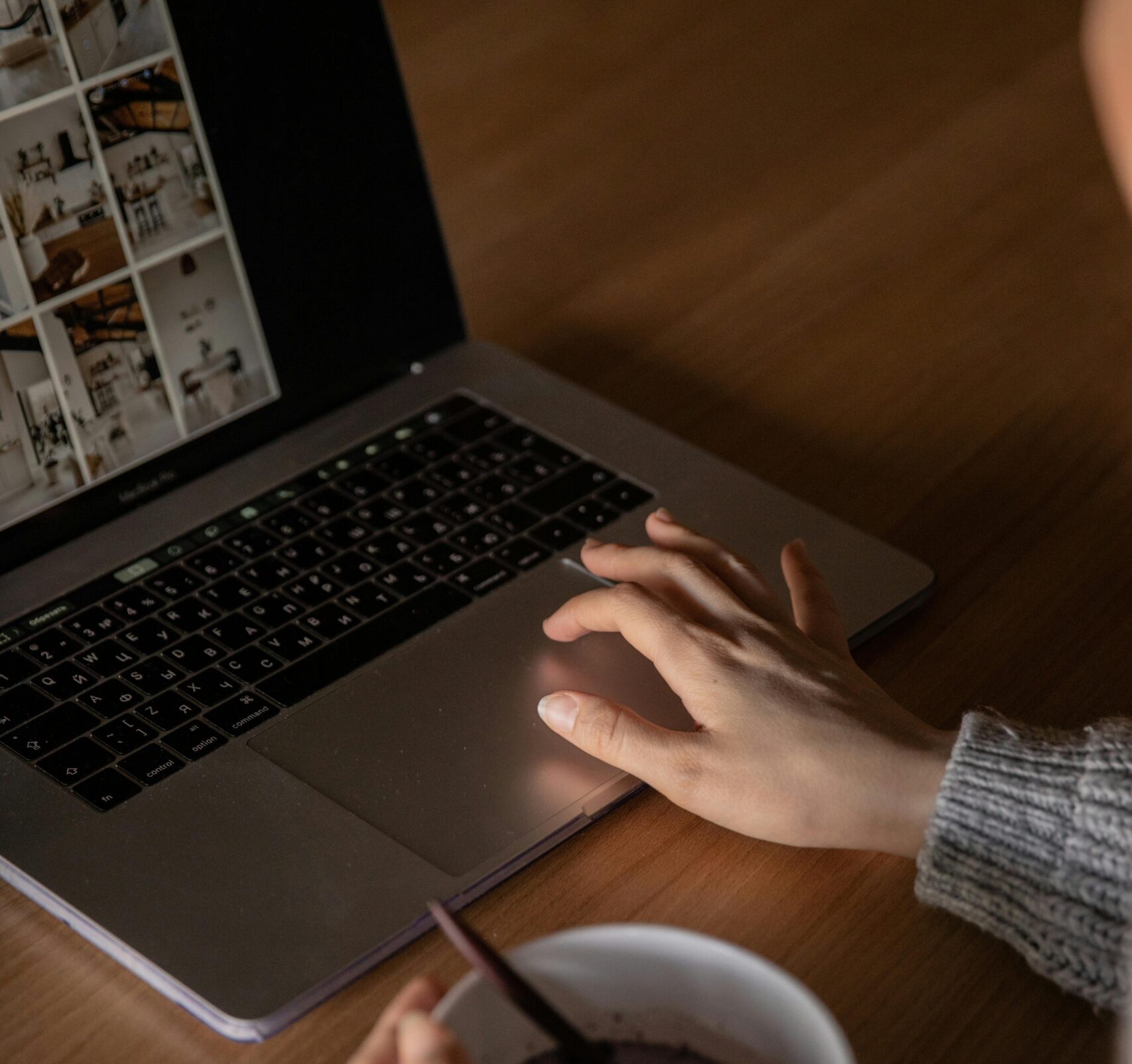 A laptop placed on a table, with a person's hand on the mouse pad. The person is also holding a cup of tea in their other hand
