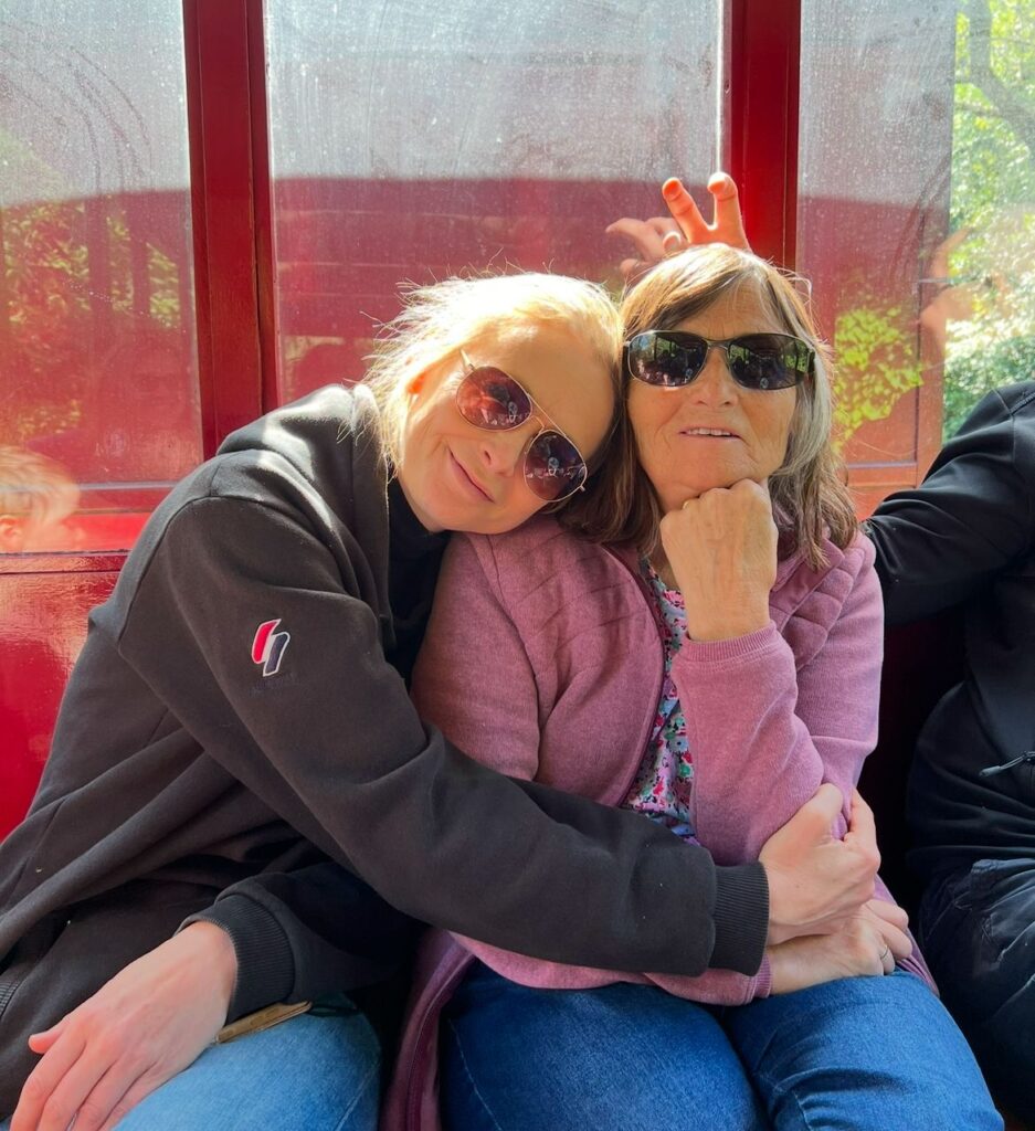 Two women sat outside together. One leans on the other's shoulder. It is sunny and they are both wearing sunglasses