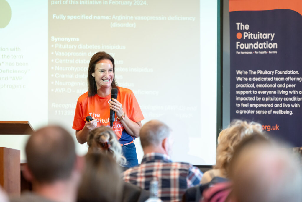 A woman is stood infront of a presentation screen, wearing an orange Pituitary Foundation t-shirt and holding a microphone. There is an audience in front of her.