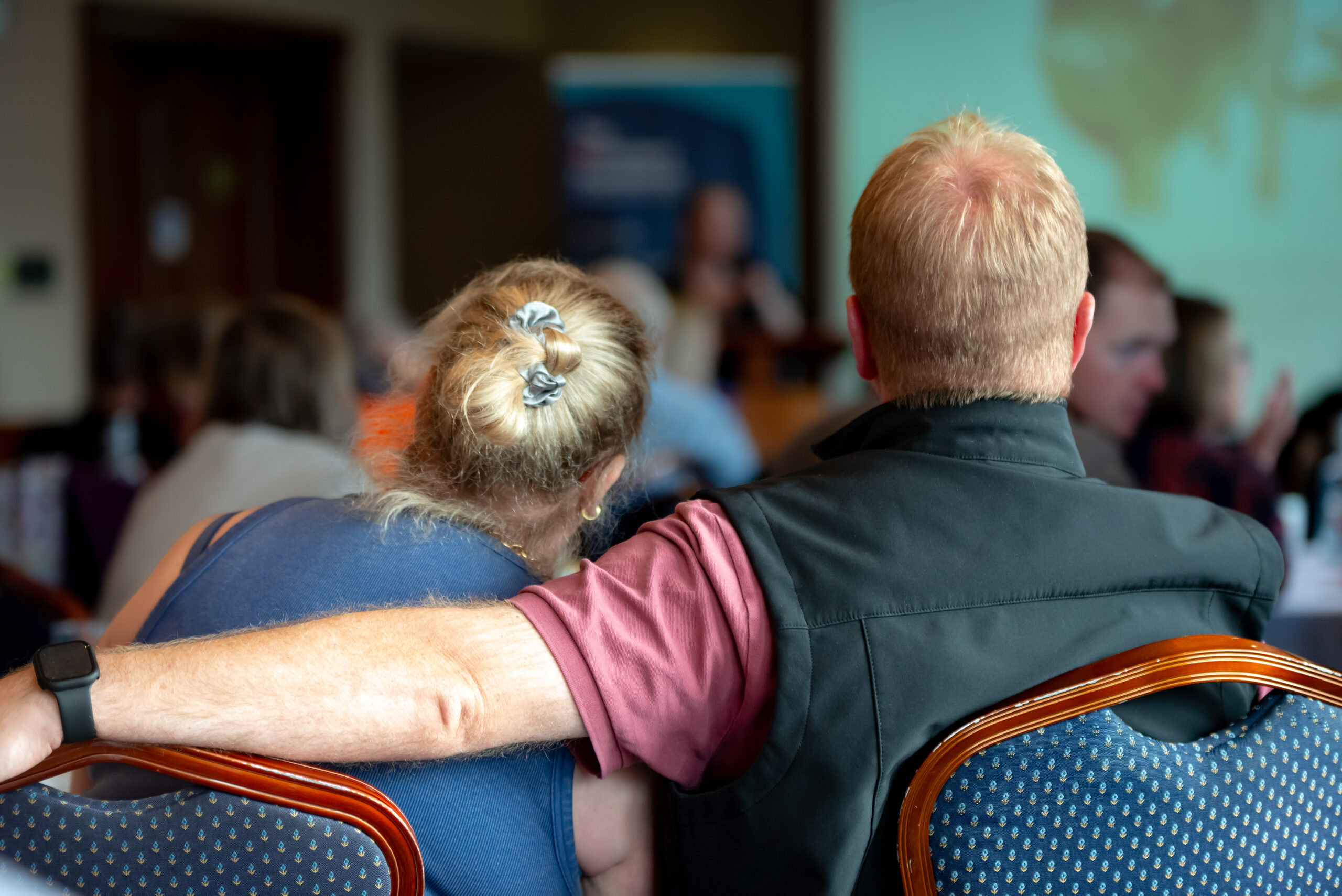A man and woman sat with their backs to the camera. The woman is leaning in towards the man's shoulder. The man has his arm around the woman.