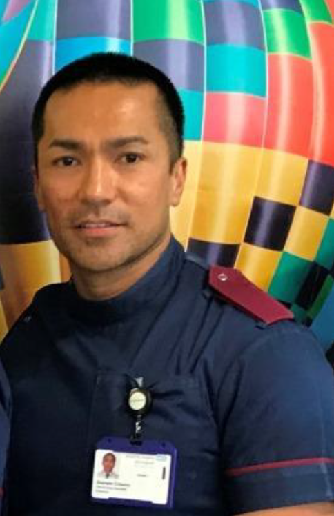 A young man in healthcare uniform is stood infront of a colour background