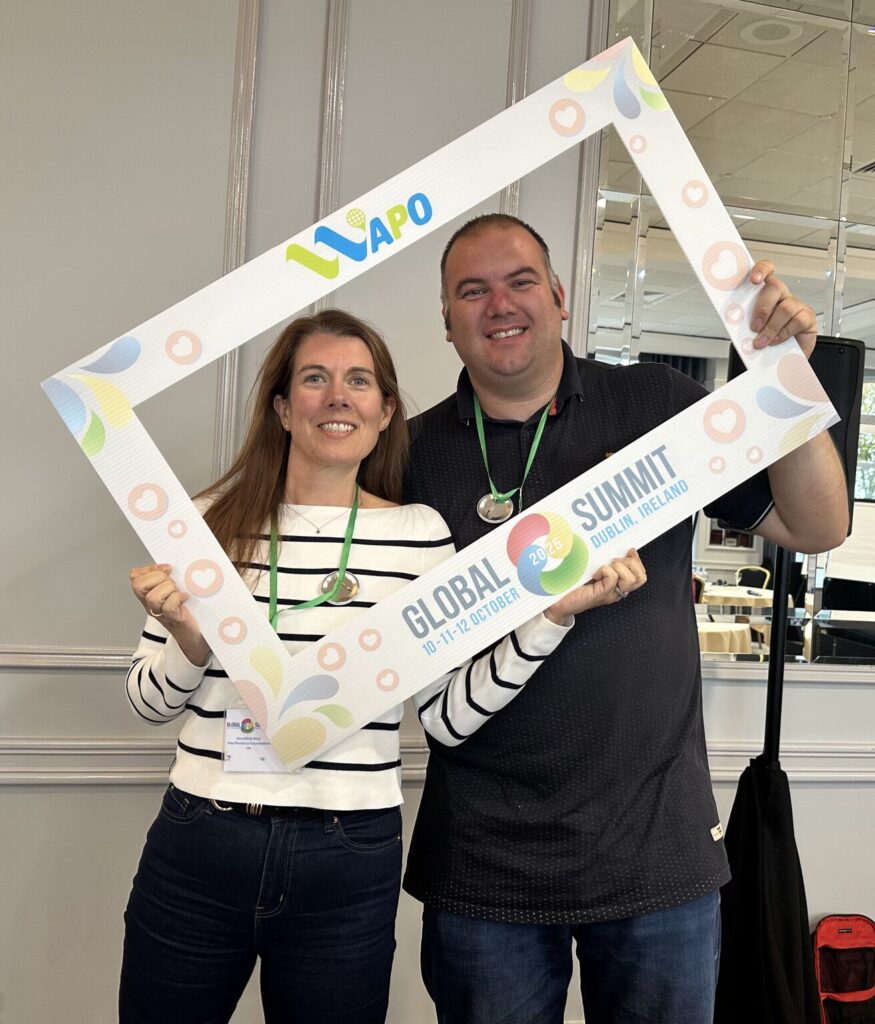 Two people stand together holding up a square cardboard photo frame adorned with WAPO branding and colours. They are smiling through the square frame.
