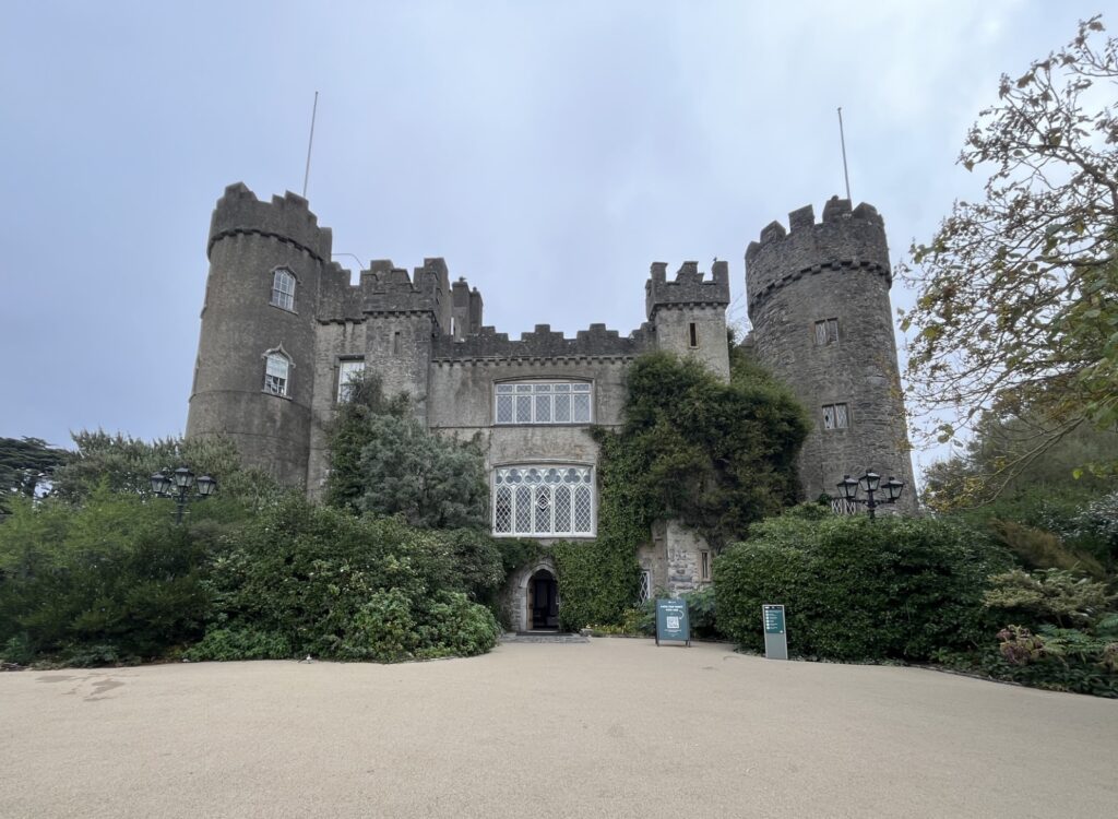 A stone castle stands against a grey, cloudy sky. Trees and shrubs surround the base of the castle, with some climbing up the walls.