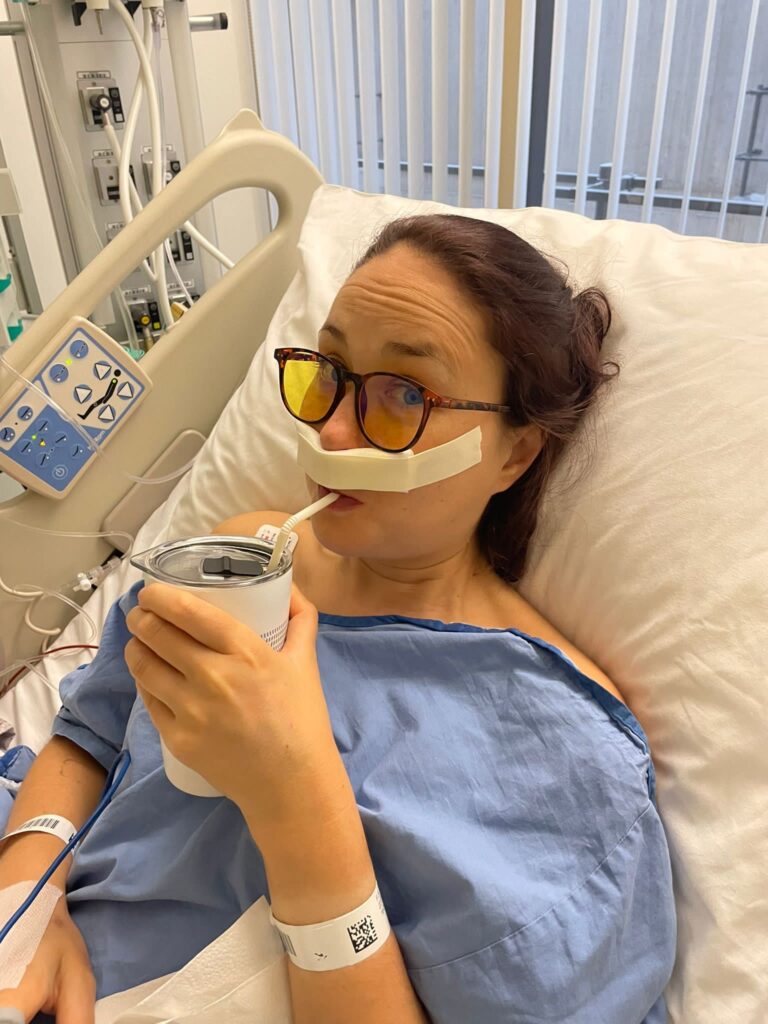 Francesca is in a hospital bed drinking from a cup out of a straw. She has medical tape and packing over her nose.