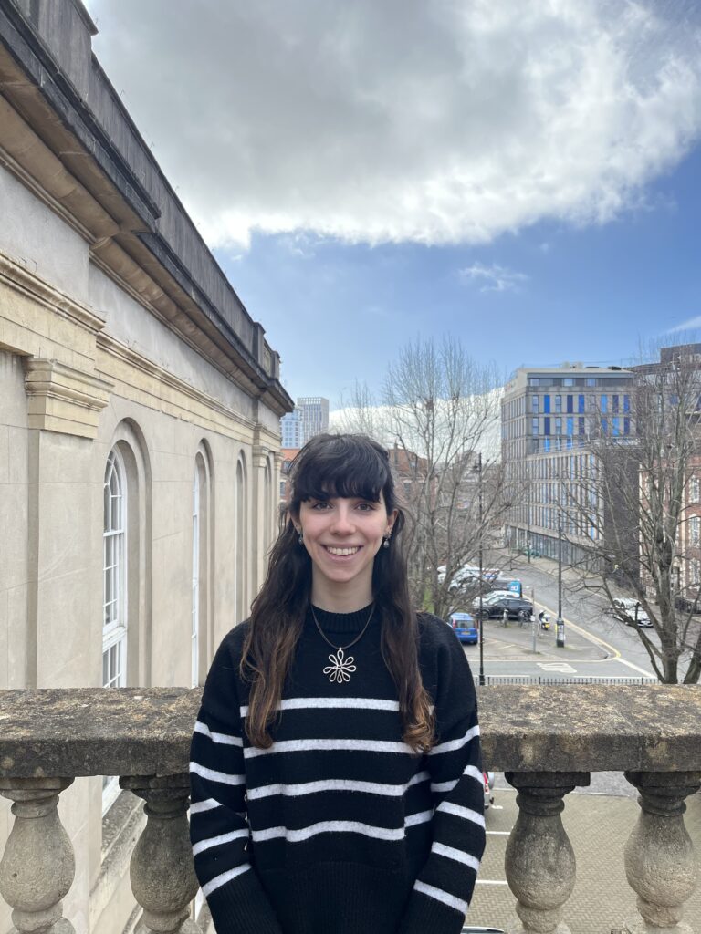 A young woman with dark hair is stood outside. She is smiling and wearing a black and white stripey jumper