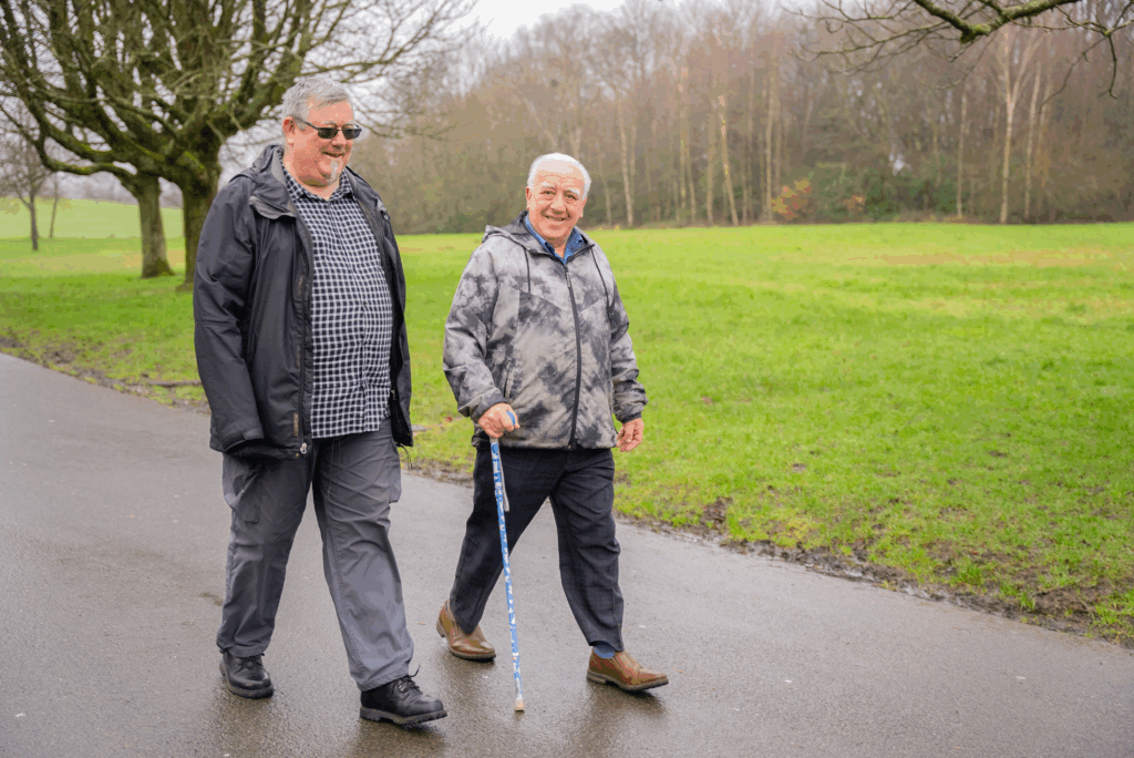 two older men out for a walk on a rainy, grey day. One is wearing dark glasses and the other has a walking stick. They are smiling. There is grass behind them