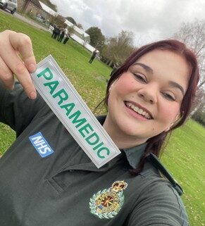 Maria wearing her paramedic uniform, holding a badge that says 'paramedic'