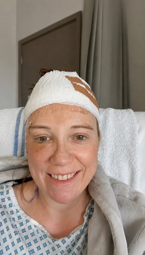 Helen smiling in her hospital gown,  sitting up with a bandage wrapped around her head. 