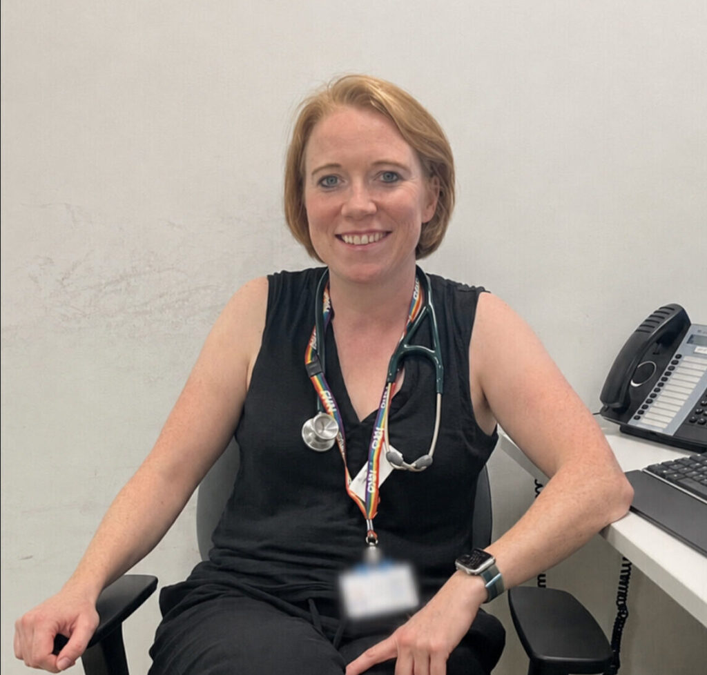 Helen wearing her NHS badges and a stethoscope, smiling and sitting in an office chair
